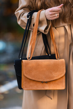 Closeup of beige trench coat draped over shoulder with tan leather handbag, black carry bag, long straps, metallic hardware, soft suede textures, urban street style details
