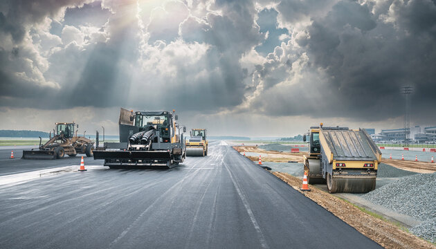 Industrial road construction machinery and asphalt paving equipment resurfacing an airport runway under a dramatic sky with sunbeams.
