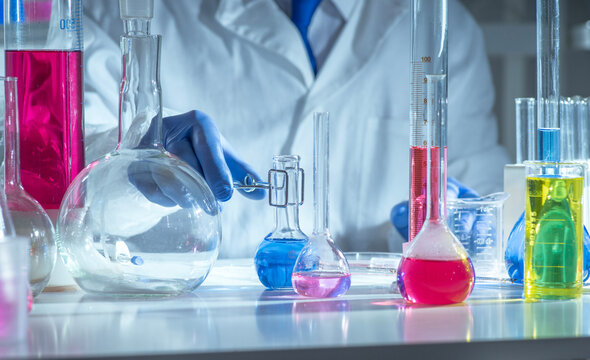 Chemistry laboratory scene with colorful chemical solutions in glass flasks and cylinders, scientist in gloves writing results, analytical experiment workspace.