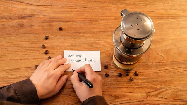 Hands writing a recipe for Vietnamese drip coffee with condensed milk on a wooden table next to a phin filter and coffee beans