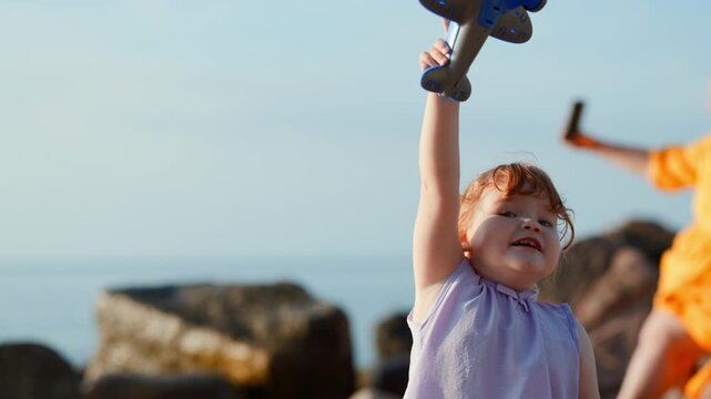 Little girl playing with toy airplane against blue sky and sea background. Child imagining flight near shore. Dream, freedom and childhood innocence. Travel and adventure concept.