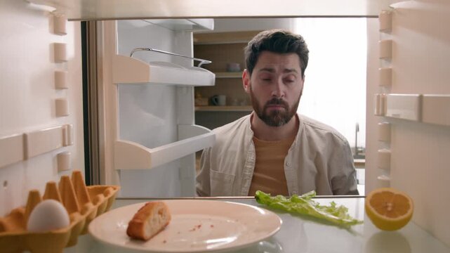 Hungry sad Caucasian man opening empty fridge half lemon lettuce leaf one egg eating bread crumbs adult male guy food groceries ordering delivery unemployment crisis in kitchen POV inside refrigerator