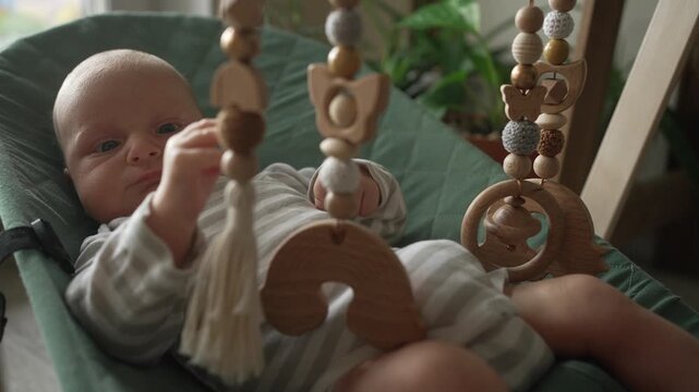 Sweet newborn infant lying in a modern green bouncer. Observing and touching a hanging wooden mobile with eco friendly toys. This moment represents early childhood development and quiet play at home