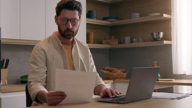 Focused Caucasian business man paying bills online laptop paperwork in kitchen male businessman checking finances mortgage payments budget documents homeowner reviewing banking planning website indoor