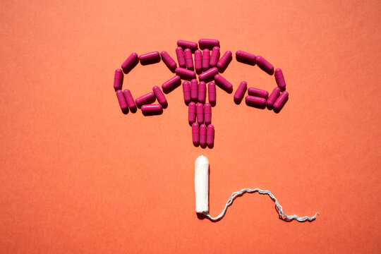 Artistic arrangement of purple capsules and a white tampon forming a uterus shape on a textured orange background, symbolizing women's health and menstrual care