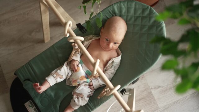 Curious newborn baby in pajamas actively engaging with a wooden play arch, lying comfortably in a modern green bouncer and exploring the hanging toys with focused attention