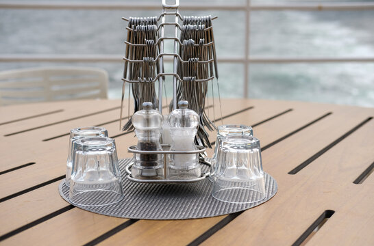 Metallbesteck mit Gew&uuml;rzstreuern und Wassergl&auml;sern auf einem gedeckten Tisch mit Blick auf das Wasser im verschwommenen Hintergrund