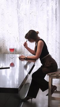 Vertical video. Female journaling. Quiet planning. Serious expert writing in notebook at table with red drink in glass using phone indoor.