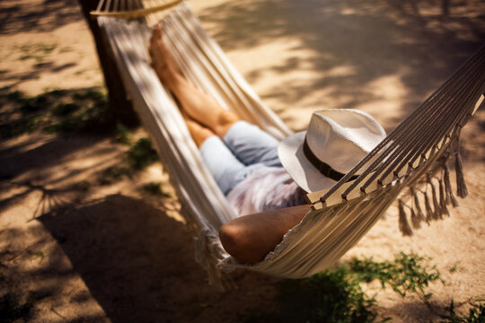 Person relaxing in a hammock wearing a wide-brimmed hat, with bare feet visible, surrounded by sandy ground and dappled sunlight filtering through trees