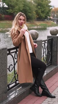 Vertical video. Autumn portrait. Outdoor rest. Woman in beige coat boots standing near park river fence touching face with tree leaves.