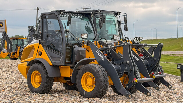 Regina, Canada - July 20, 2026: Two John Deere compact wheel loaders displayed at dealership lot on gravel with backhoe in background