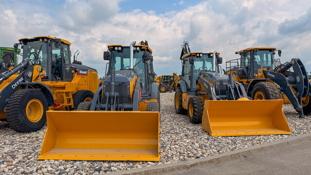 Regina, Canada - July 20, 2026: Row of John Deere yellow wheel loaders and backhoe loaders parked on gravel lot at Brandt dealership under cloudy sky