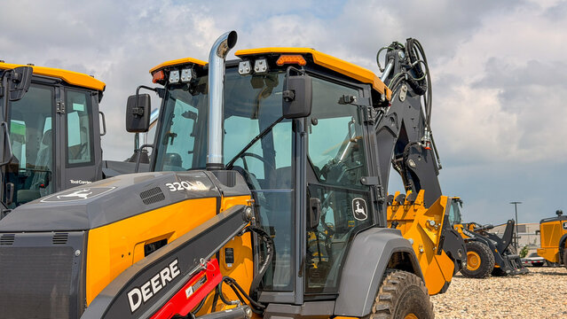 Regina, Canada - July 20, 2026: John Deere 320L backhoe loader on dealer lot with hydraulic arm raised against cloudy sky
