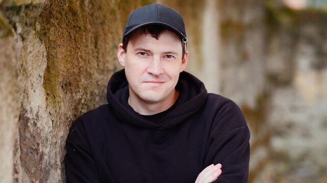 Young man in black hoodie and baseball cap leaning against textured stone wall. Urban portrait of a guy looking at camera. Casual lifestyle session for fashion.