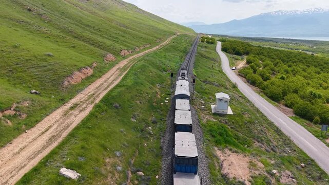 The camera tracks directly above a moving freight train, following its route through green countryside with apricot orchards, a mountain, a parallel road and distant snow-capped peaks.
