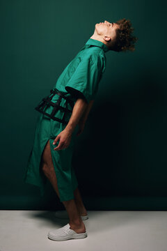 Man with curly hair wearing oversized green pareo skirt with large slit and green shirt with wide sleeves bending backwards against plain studio green background.