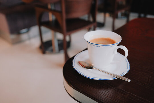White porcelain coffee cup with saucer and spoon sits on a dark wooden cafe table in a quiet modern interior