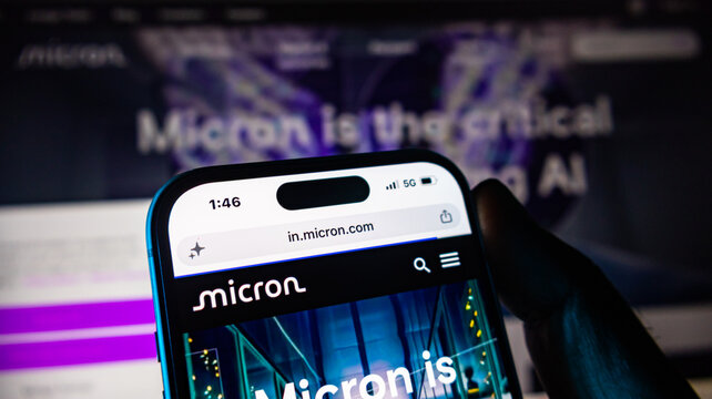A close up of a hand holding an iphone 14 pro displaying micron.com website on its screen.