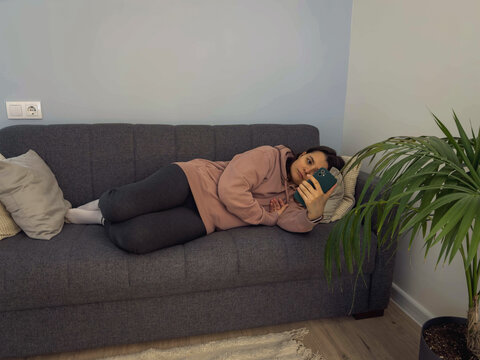 Young brunette woman lying on sofa absorbed by smartphone screen illustrating phone addiction compulsive scrolling procrastination digital dependence modern home lifestyle technology concept