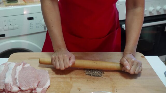 Person using a rolling pin to flatten meat, in kitchen, with rolling pin, meat, cutting board, from a close-up slightly above eye level perspective, with red, brown accents, and concentrated mood.