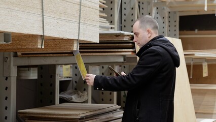 Skilled carpenter comparing lumber prices on smartphone, holding particle board sample in hardware store warehouse, evaluating material options for construction project © Alexander