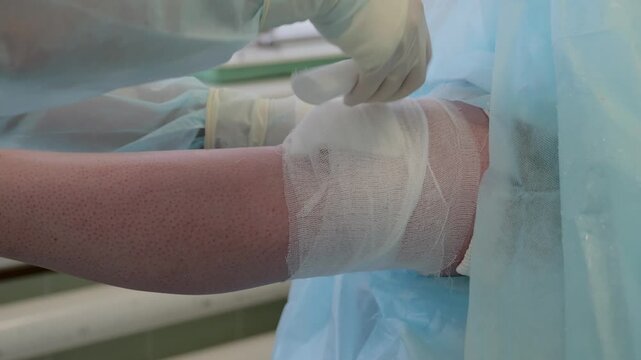 Gloved medical worker securing sterile gauze and adhesive tape over intravenous catheter on patient forearm during clinical care procedure in bright treatment room