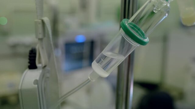Close view of intravenous infusion drip chamber with clear tubing, green cap, stainless pole, and blurred clinical ward background during saline delivery in sterile healthcare setting