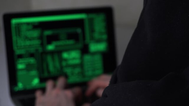 A man in a hoodie types on a laptop with a green screen glow. The man stares at the laptop intently while green light from the screen highlights his hands.