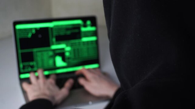 A man in a hoodie types on a laptop with a green screen glow. The man stares at the laptop intently while green light from the screen highlights his hands.
