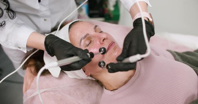 Clinic procedure. Facial therapy. Female beauty master in black gloves using professional microcurrent device for skin rejuvenation.