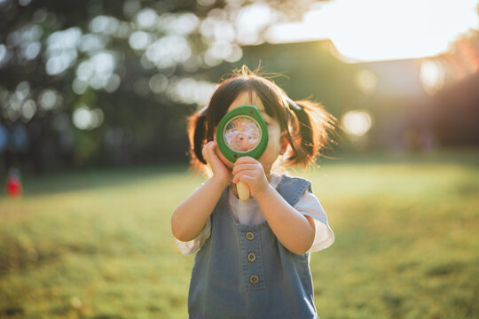 Curious Asian toddler girl looking through a magnifying glass in a sunny garden. Childhood discovery exploration and outdoor learning concept. Little explorer playing in the park during golden hour