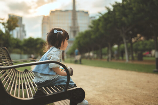 Little Asian toddler girl sitting on park bench holding toy during sunset, cute young kid enjoying family travel vacation outdoors near famous landmark for childhood, tourism and lifestyle concept.
