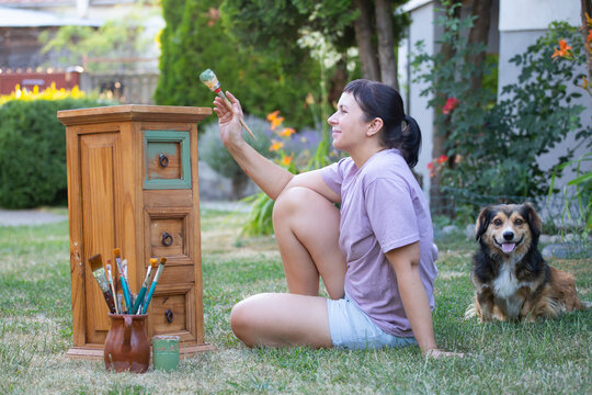 Woman painting a wooden furniture while sitting with her dog in the garden. DIY concept.
