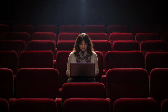 Mid adult Caucasian woman working on a laptop in an empty cinema auditorium under a single spotlight, creative professional or scriptwriter in a theater