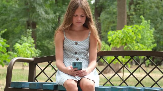 Preteen girl sitting alone on bench in city park and texting on her phone. Kid using smartphone outdoors