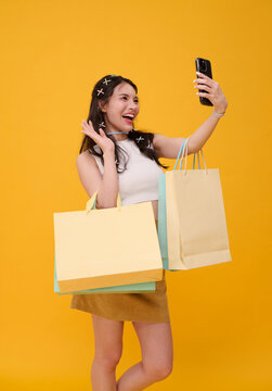 Shopping selfie with joyful Asian woman holding colourful bags and smartphone in studio expressing retail excitement social sharing consumer confidence and modern mobile lifestyle.