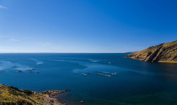 Wide scenic view over Lake Titicaca from a rocky shoreline, showing floating aquaculture trout farms dotting the deep blue water with the distant snow-capped Cordillera Real mountains on the horizon