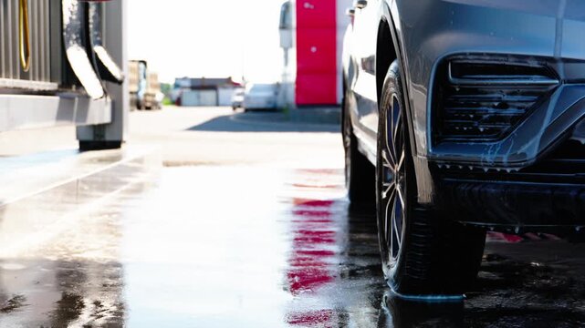 Wet pavement reflects red and white hues near a car. Car wash nozzles spray water over a dark SUV's front bumper. White sedan waits behind the vehicle. Scene shows routine vehicle cleaning