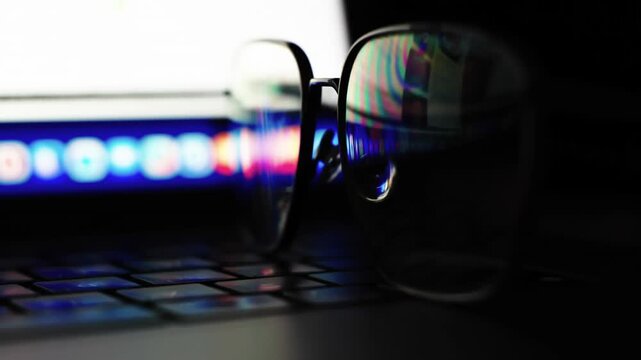 Glasses rest on keyboard, reflecting glowing screen. Darkness frames a focused, digital workspace. Blue light pulses softly beneath the lenses. Silent, still moment before the next keystroke