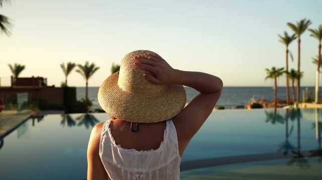 Woman in straw hat stands poolside at sunset. She waves, gazing at distant ocean horizon. Palm trees line infinity pool edge. Warm golden light bathes tranquil resort scene. Perfect for vacation