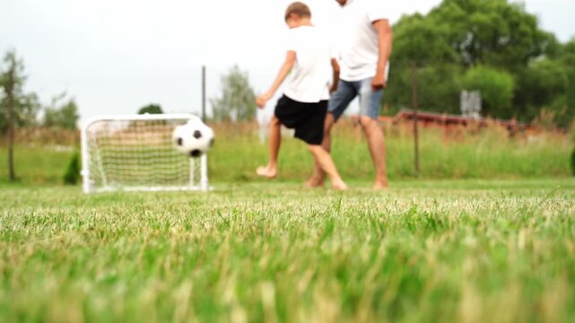 Sunlit grass field with two figures playing soccer. One child runs after a ball near a small goal. Adult watches nearby, encouraging the child. Joyful, carefree motion of outdoor family fun