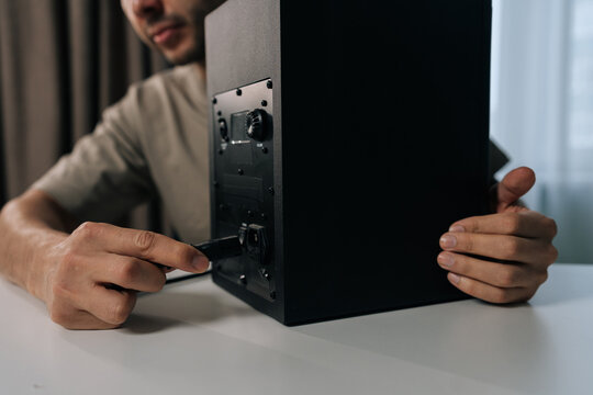 Cropped shot of young man inserting power cord into back of black studio monitor speaker, ensuring secure connection for audio setup on clean white table.
