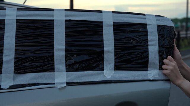 Female hands using white adhesive tape to attach black plastic bag over broken car window, providing temporary fix for damaged automobile glass after accident, vandalism, close-up, slow motion.