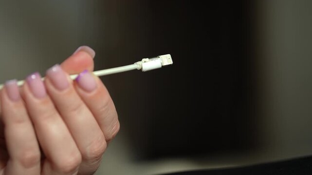 Woman hand with manicure holding and inspecting broken white phone charging cable. Close-up cropped shot of frayed wire and damaged connector, concept of planned obsolescence and e-waste, slow motion.