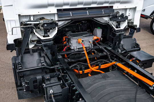 Electric truck chassis frame with inverter unit and battery pack. EV commercial transport vehicle powertrain mechanism under white cab with high voltage orange cables engineering details.