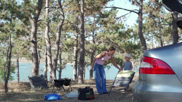 A caring mother helps her joyful son open a folding chair in a sunny pine forest. They feel happy setting up a camp near the blue lake. The peaceful woods feel warm and relaxing.