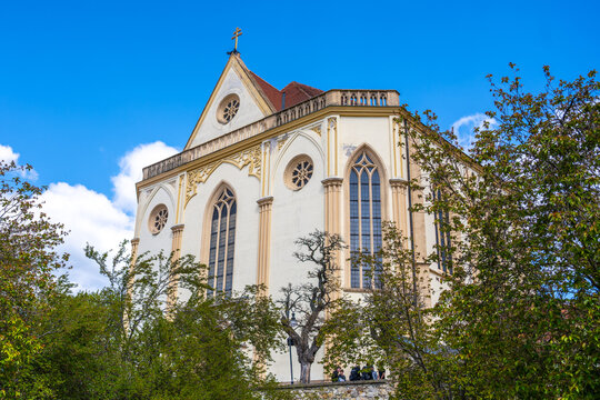 Visitors gather near the Church of St. James the Elder in Boskovice, Czechia. The building shows its architecture against a clear blue sky with some clouds.
