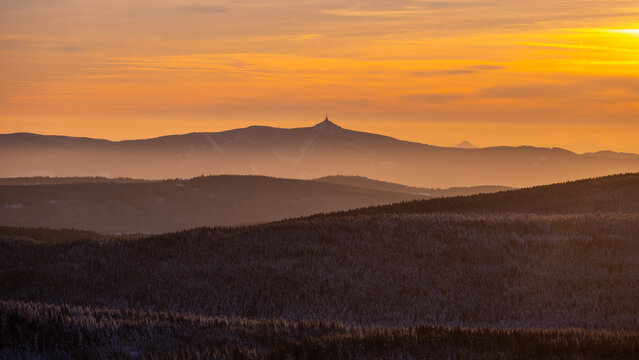 The sun sets behind the Jested mountain ridge. The sky displays various colors as the light fades. Shadows stretch over the landscape, revealing the silhouette of the ridge.