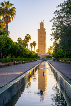 The Koutoubia minaret rises above the trees in Marrakesh. The sun sets, casting a warm light on the building. A calm water channel reflects the tower and the surrounding gardens.