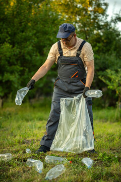 A worker collects plastic bottles and rubbish in a green field. He wears gloves and works to clean the area. The sun shines through trees in the background